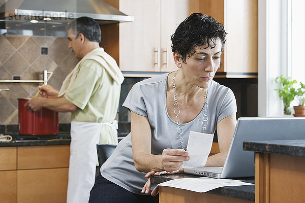 Ein Mann kocht das Abendessen an der Herdplatte  und eine Frau benutzt einen Laptop und überprüft den Papierkram in der Küche.