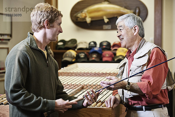 Asiatisch-amerikanischer älterer männlicher Besitzer eines Fliegenfischer-Einzelhandelsladens im Gespräch mit einem jungen kaukasischen Mann über die Vorzüge der neuen 5-Gewicht-Fliegenfischerrute. Der Besitzer trägt eine Fliegenfischerweste.