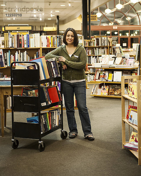 Porträt einer kaukasischen Mitarbeiterin  die in einer Buchhandlung neben einem Bücherwagen steht.