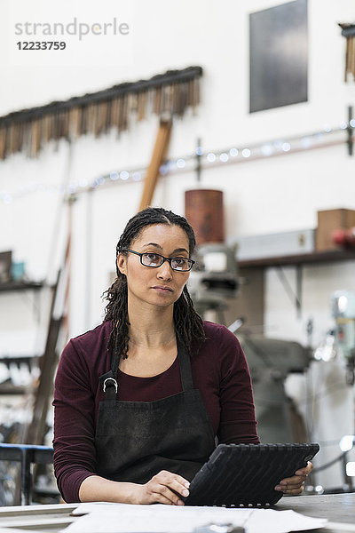 Frau mit Brille und Schürze steht an der Werkbank in einer Metallwerkstatt  hält ein digitales Tablett in der Hand und schaut in die Kamera.