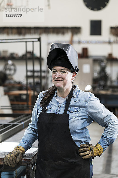 Porträt einer Frau mit Schürze und Schweißermaske  die in einer Metallwerkstatt steht und in die Kamera lächelt.