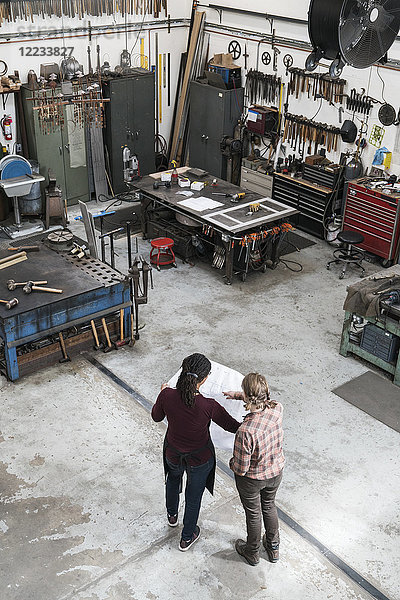 Hochwinkelaufnahme von zwei Frauen  die in einer Metallwerkstatt stehen und einen technischen Bauplan in der Hand halten.