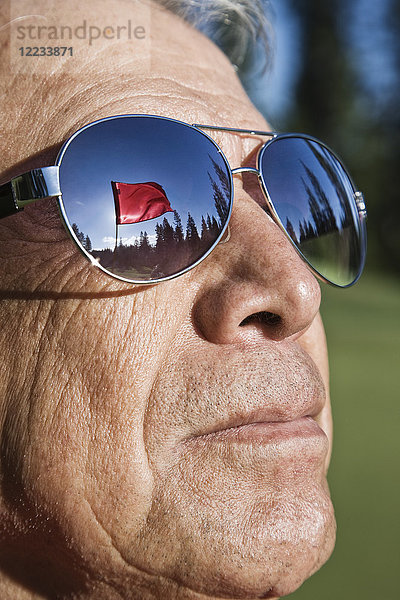 Eine Golfflagge auf einem Grün  die sich in der Sonnenbrille eines älteren Golfspielers spiegelt.