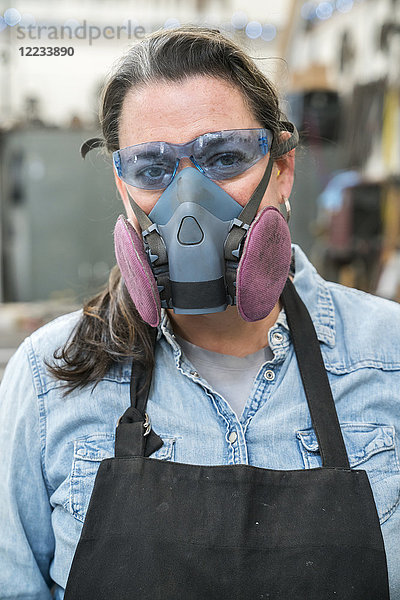 Porträt einer Frau mit Schutzbrille und Staubmaske  die in einer Metallwerkstatt steht und in die Kamera schaut.