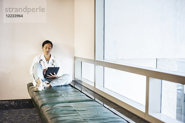 Asiatische Ärztin  die einen Notebook-Computer benutzt und einen Laborkittel und ein Stethoskop trägt.
