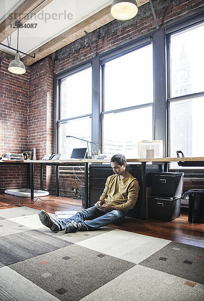Ein spanischer Mann mit seinem Mobiltelefon  während er an seinem Büroarbeitsplatz auf dem Boden sitzt.