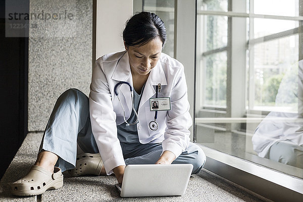 Asiatische Ärztin  die auf einem Schoß-Top in einem Krankenhausflur arbeitet.