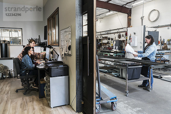 Zwei Frauen an einem Schreibtisch im Bürobereich einer Metallwerkstatt  Frau steht an einer mobilen Werkbank.
