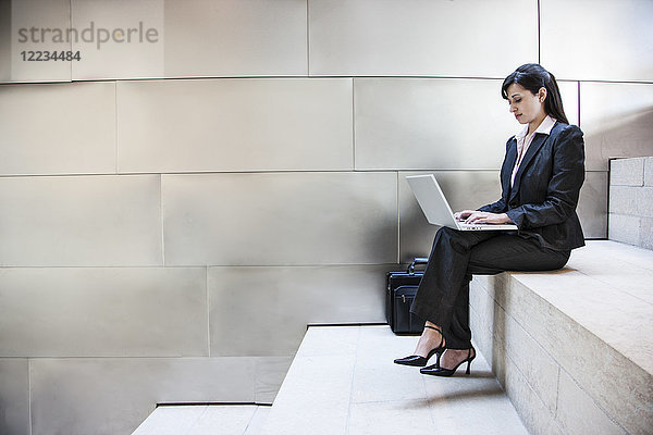 Eine hispanische Geschäftsfrau sitzt auf einer Treppe in der Lobby eines großen Bürogebäudes.