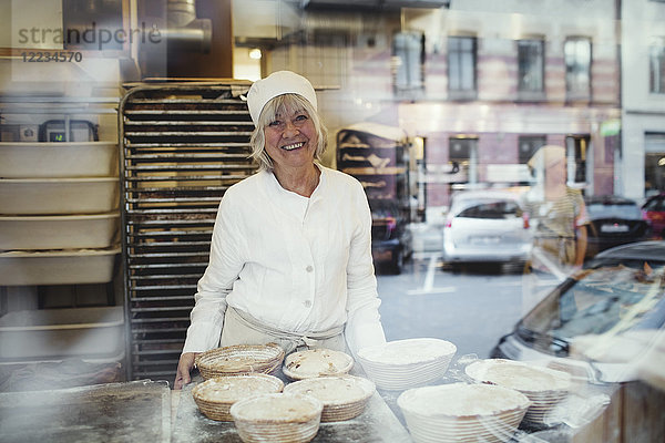 Porträt des ältesten lächelnden Bäckers in der Bäckerei vom Glasfenster aus gesehen