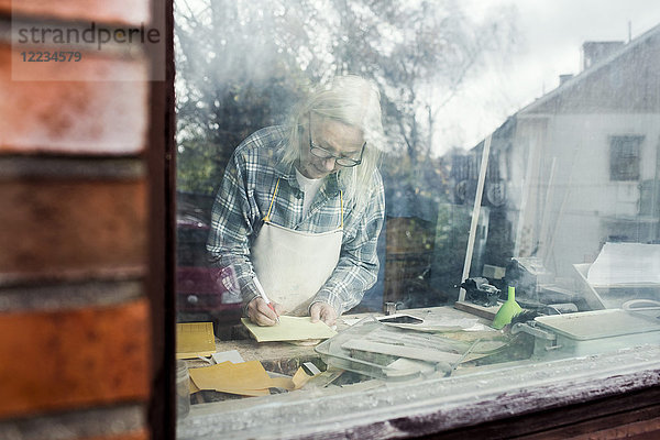 Senior Handwerker beim Schreiben auf Papier durch Glasfenster gesehen