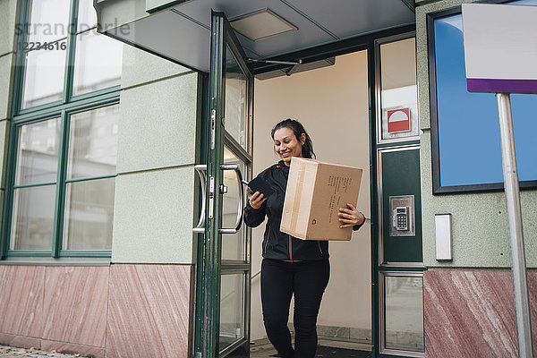 Lächelnde Arbeiterin mit Box mit Handy beim Verlassen des Gebäudes