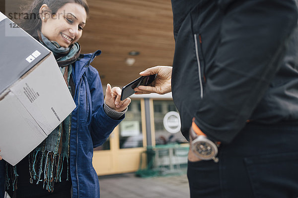 Lächelnde Frau benutzt Smartphone  um für ihre Lieferung vom Boten zu unterschreiben.