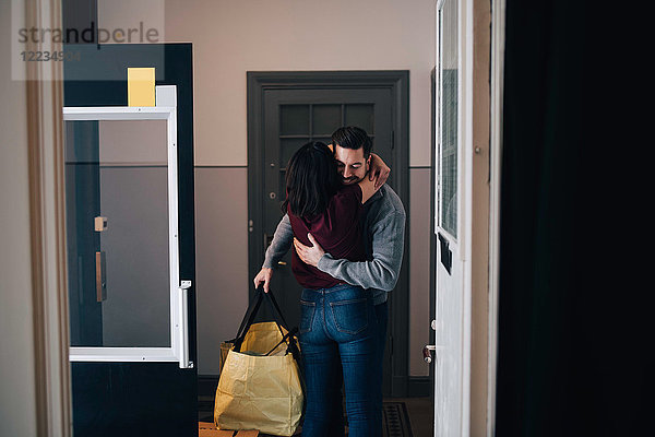 Frau umarmt den Mann mit Tasche  während sie im Flur steht  von der Tür aus gesehen.