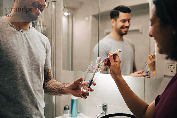 Lächelndes Paar  das Zahnbürsten in einem Behälter mit Spiegel im Badezimmer anordnet.