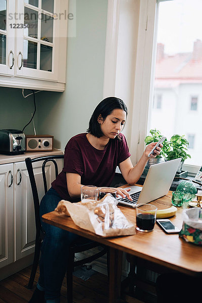 Frau benutzt Laptop  während sie das Handy am Esstisch in der Küche hält.