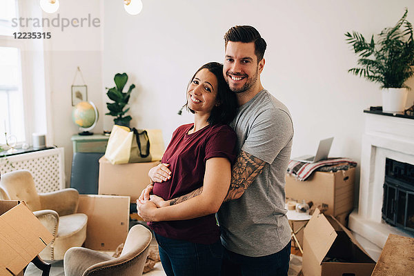 Lächelnder Mann  der schwangere Frau umarmt  während er im Wohnzimmer gegen Kisten steht.