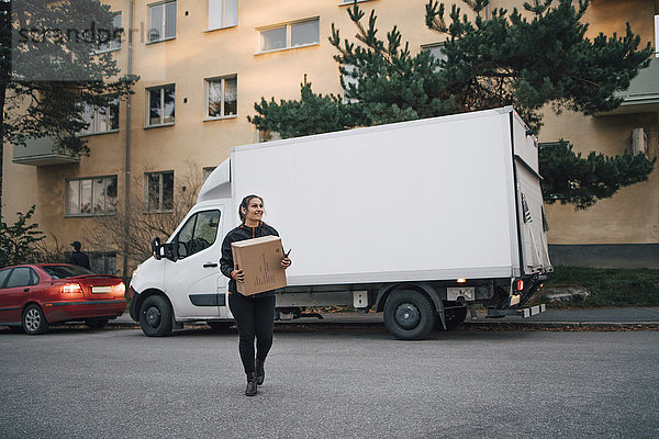 Volle Länge der Arbeiterin  die die Kiste trägt  während sie auf der Straße gegen den Lieferwagen läuft.