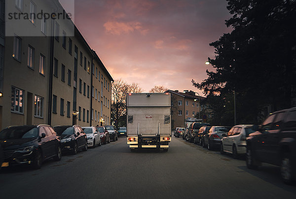 Lieferwagen auf der Straße inmitten geparkter Autos in der Stadt gegen den Himmel bei Sonnenuntergang