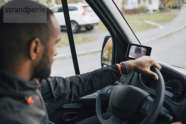 Männlicher Arbeiter schaut auf den Bildschirm des Geräts  während er einen Lieferwagen in der Stadt fährt.