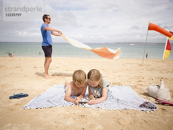 Mann hält Handtuch  während Kinder sich am Strand ein Smartphone gegen den Himmel teilen.