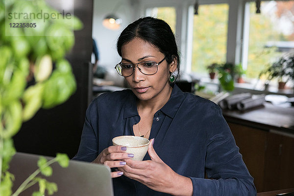 Geschäftsfrau hält Tasse beim Blick auf Laptop im Home-Office