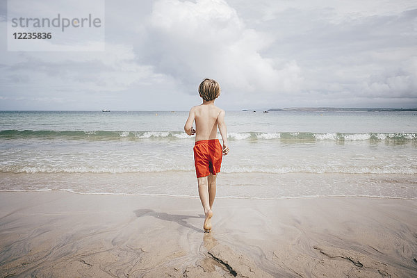 Rückansicht des shirtlosen Jungen  der am Strand gegen den Himmel läuft.