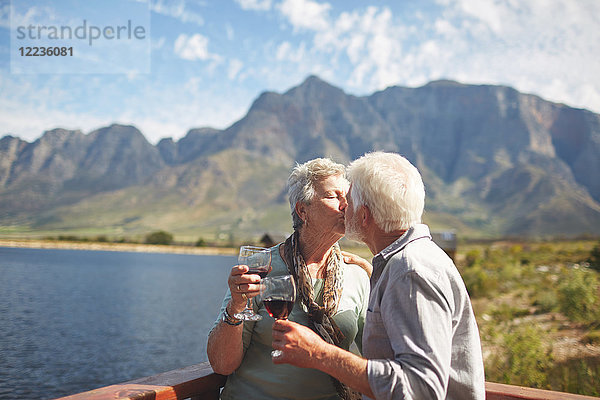 Romantisches  aktives Seniorenpaar küsst und trinkt Wein auf dem sonnigen Sommerbalkon am Seeufer.