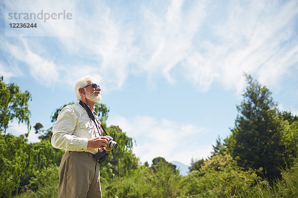 Neugieriger aktiver Senior mit Digitalkamera und Blick auf sonnige Bäume und Himmel