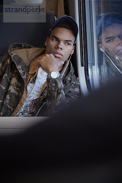 Seriöser  nachdenklicher junger Mann  der im Personenzug aus dem Fenster schaut und mit Kopfhörern Musik hört.
