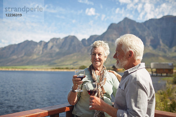 Lächelndes aktives Seniorenpaar trinkt Rotwein auf dem sonnigen Sommerseebalkon