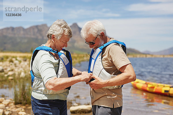 Aktives Seniorenpaar beim Anziehen von Schwimmwesten  Vorbereitung auf das Kajakfahren am sonnigen Sommerseeufer