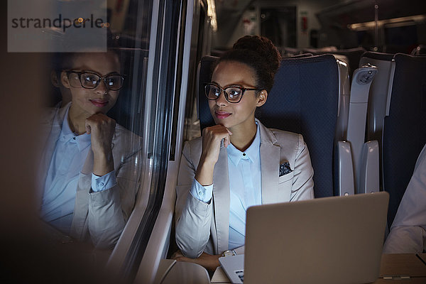 Selbstbewusste  nachdenkliche Geschäftsfrau  die nachts im Personenzug aus dem Fenster schaut und am Laptop arbeitet.