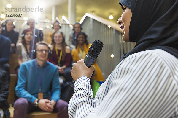 Lächelnde Geschäftsfrau im Hijab  die mit Mikrofon vor Publikum spricht.