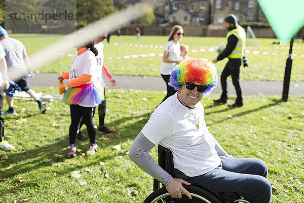 Portrait lächelnder  verspielter Mann im Rollstuhl mit Clown-Perücke beim Charity-Rennen im sonnigen Park