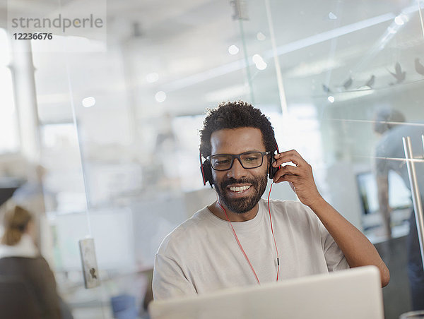 Lächelnder kreativer Geschäftsmann mit Kopfhörern bei der Arbeit am Laptop im Büro