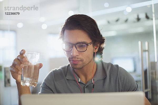 Konzentrierter Geschäftsmann mit Kopfhörern  der am Laptop Wasser trinkt