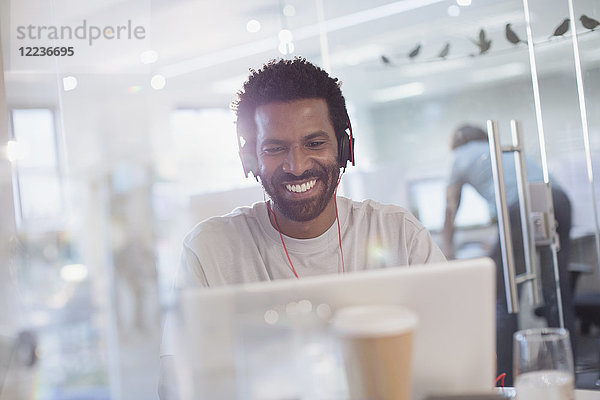 Lächelnder  selbstbewusster  kreativer Geschäftsmann mit Kopfhörern und Laptop im Büro