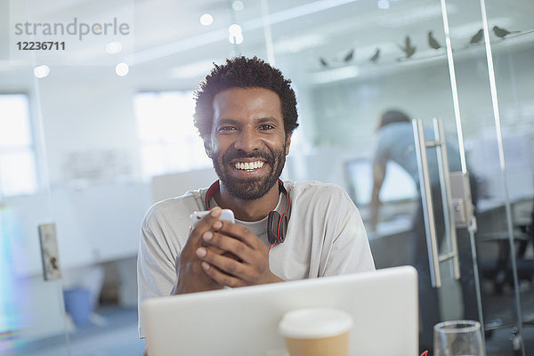 Porträt eines lächelnden  selbstbewussten  kreativen Geschäftsmannes  der ein Smartphone am Laptop im Büro benutzt