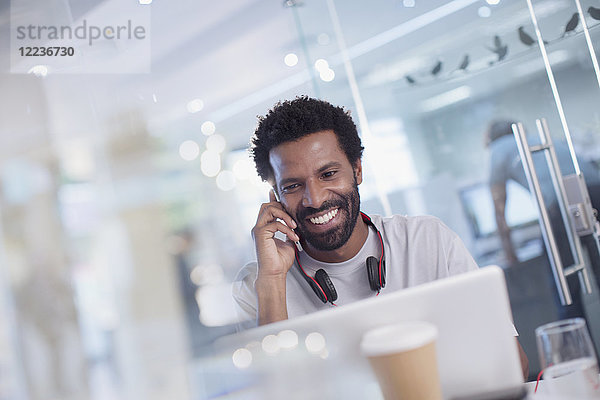 Lächelnder  selbstbewusster  kreativer Geschäftsmann  der mit seinem Smartphone am Laptop im Büro spricht