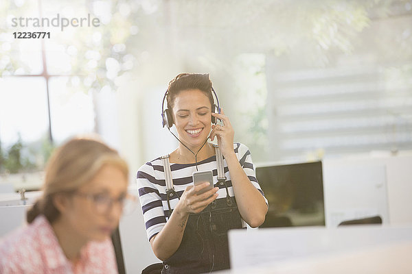 Lächelnde kreative Geschäftsfrau  die mit Kopfhörern und Smartphone Musik hört