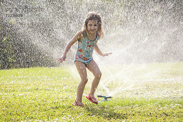 Mädchen (4-5) spritzt im Sprinklerwasser auf dem Rasen