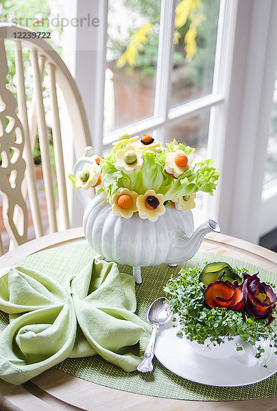 Blumenstrauß aus Salatblättern und Blumen mit Käse  Karotten und Avocado in einer Teekanne  Brunnenkresse und Karotten in einer Teetasse auf einem grünen Tischset