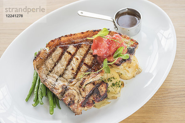 Gegrilltes Steak mit langen grünen Bohnen  Kartoffeln und gegrillten Tomaten mit Steaksauce auf einem weißen Teller und hellem Holzuntergrund