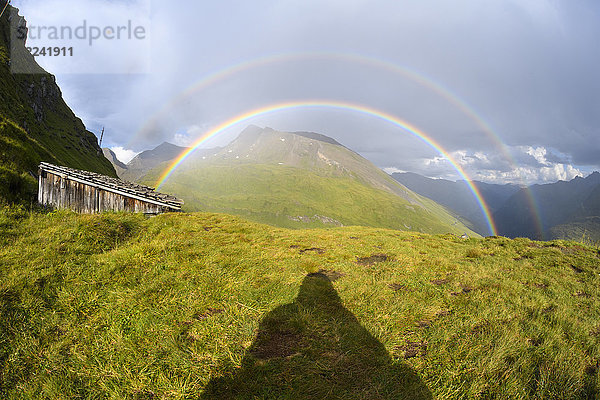 Schatten eines Mannes mit Blick auf einen Regenbogen über einer Berglandschaft auf der Kaiser Franz Josefs Hohe in der Großglockner Hochalpenstraße im Nationalpark Hohe Tauern  Kärnten  Österreich