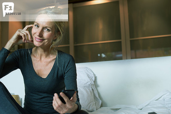 Ausgereiftes Sitzen auf dem Bett mit Smartphone in der Hand  lächelnd  Portrait