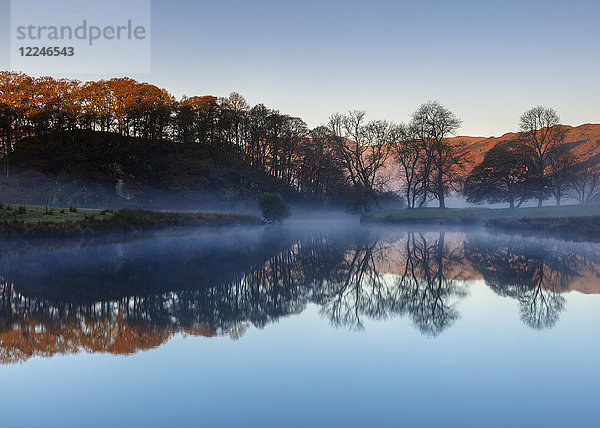 Das Licht der Morgendämmerung enthüllt die Herbstfarben der Bäume und die fernen Berge  während der Nebel über Elterwater eine perfekte Spiegelung zeigt  Lake District National Park  UNESCO-Weltkulturerbe  Cumbria  England  Vereinigtes Königreich  Europa