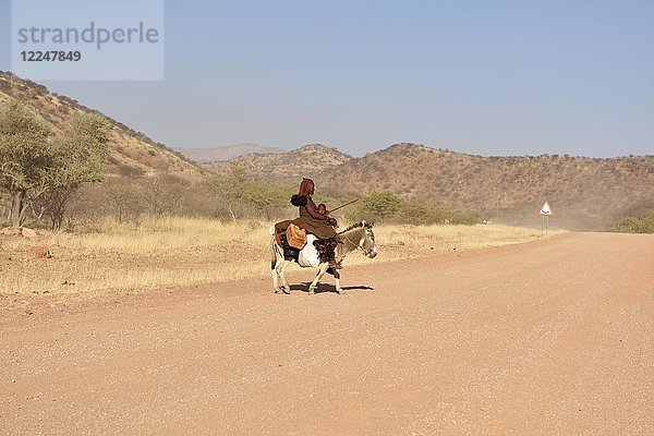 Junge  verheiratete Himbafrau reitet mit einem kleinen Kind auf einem Esel  Kaokoveld  Namibia  Afrika