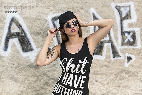 Junge tätowierte Frau im Hemd und mit Mütze vor der Graffitiwand