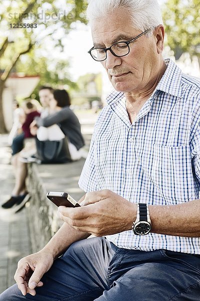 Grauhaariger Senior sitzt auf einer Mauer mit seinem Smartphone in der Hand  Köln  Nordrhein-Westfalen  Deutschland  Europa